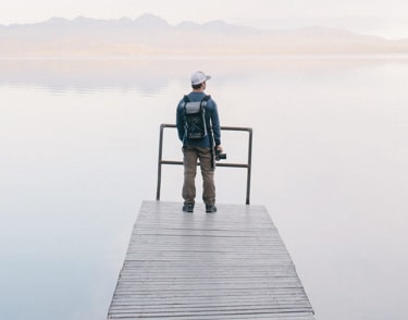 Man in a hat and backpack stands on a wooden dock overlooking a calm lake.
